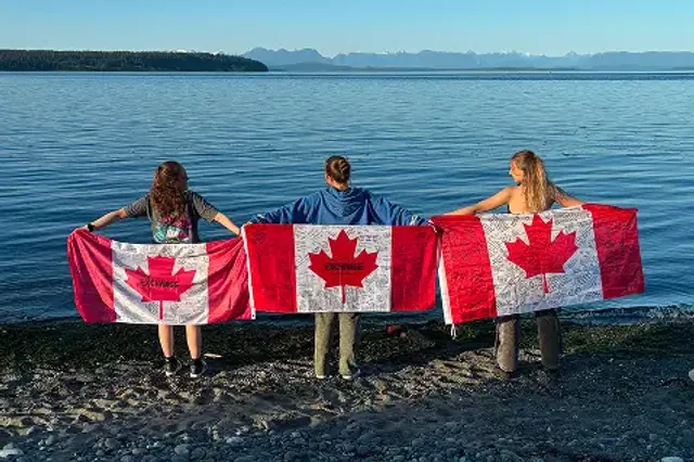 Tre studentesse di spalle reggono bandiere canadesi su una spiaggia guardando il mare