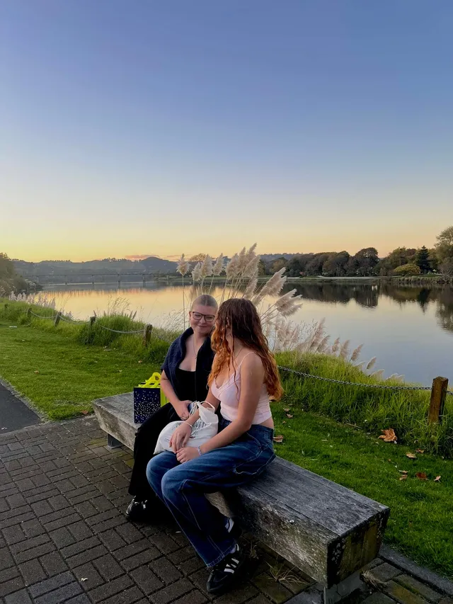 Dos chicas sentadas en un banco junto al río al atardecer, entre hierba de la pampa