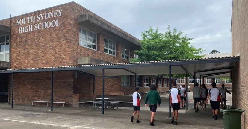 L'edificio in mattoni della South Sydney High School è circondato da alberi, mentre gli studenti passeggiano sul marciapiede.