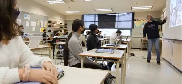 Estudiantes con mascarilla sentados en un aula mientras el profesor enseña en la pantalla