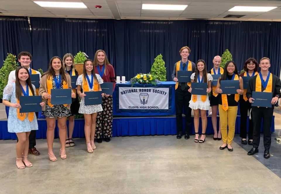 Un gruppo di giovani donne in toga da diploma sta davanti a uno striscione della National Honor Society nel campus della Clovis High School tenendo in mano i propri diplomi.