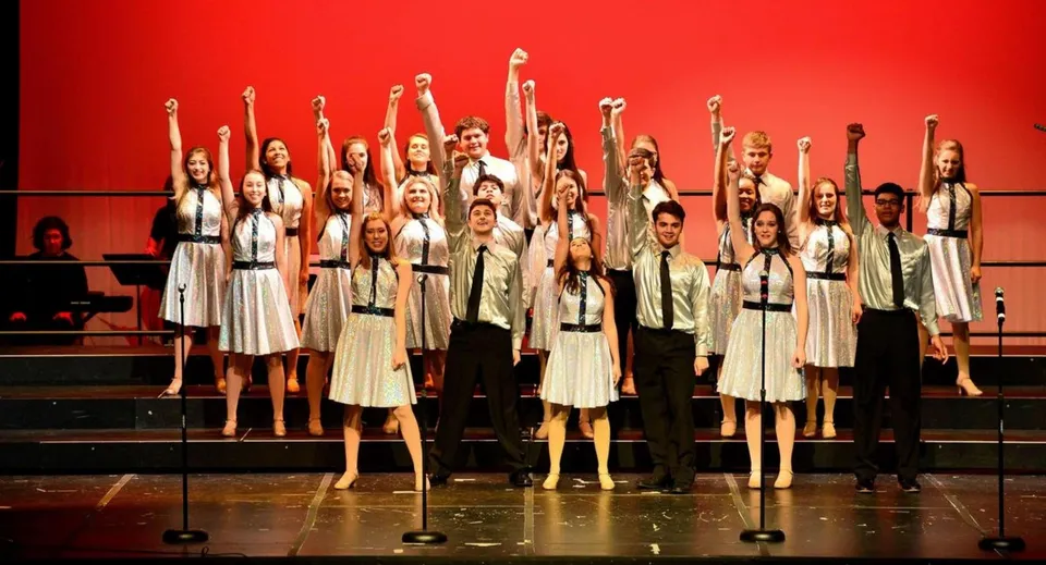 Gli studenti della Tri-State Christian School sono sul palco in outfit bianchi e alzano le braccia davanti a uno sfondo rosso.