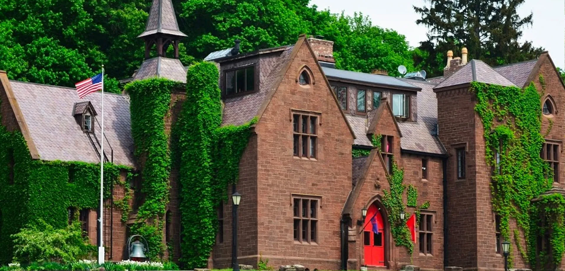 L'edificio in mattoni della Hoosac School con la porta rossa è circondato da alberi verdi e piante.