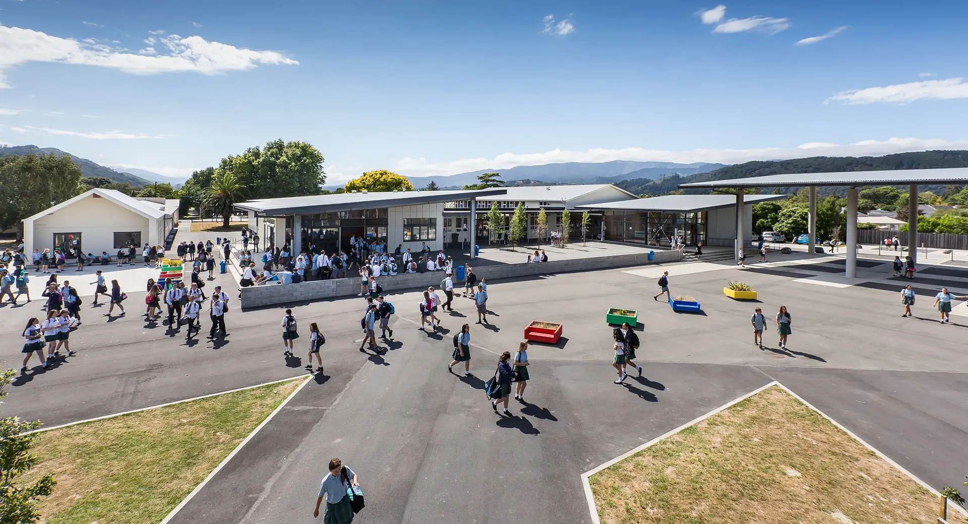 All'esterno dell'Upper Hutt College si vede una piazza del mercato animata con diversi banchi e montagne sullo sfondo.