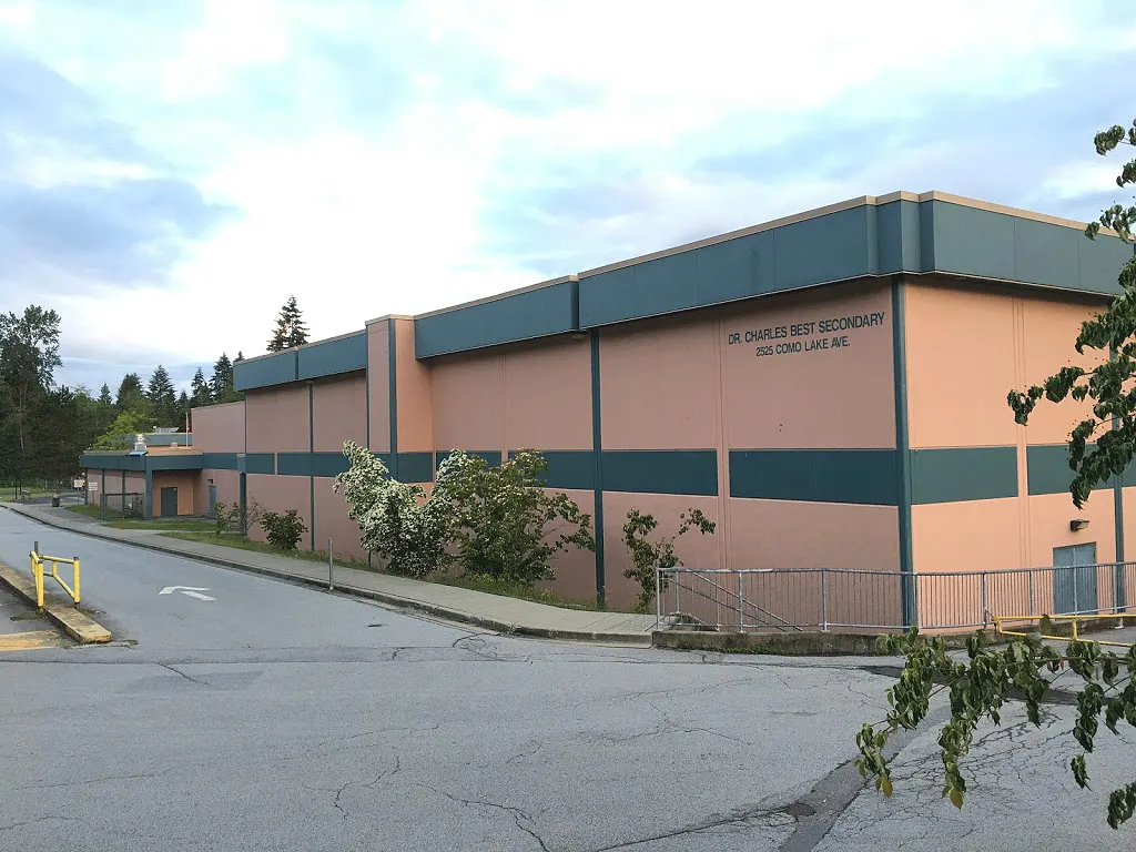 El edificio de varios pisos de la Dr. Charles Best Secondary School con fachada rosa y detalles verdes está rodeado de árboles.