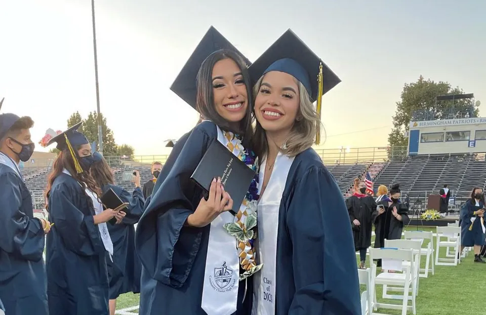 Due giovani donne in toga da diploma sono insieme davanti a uno stadio nel campus della Birmingham Community Charter High School.