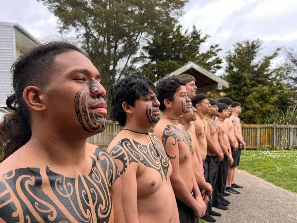 Un gruppo di studenti tatuati si trova su un prato davanti a una struttura in legno nei terreni del Nelson College for Boys.