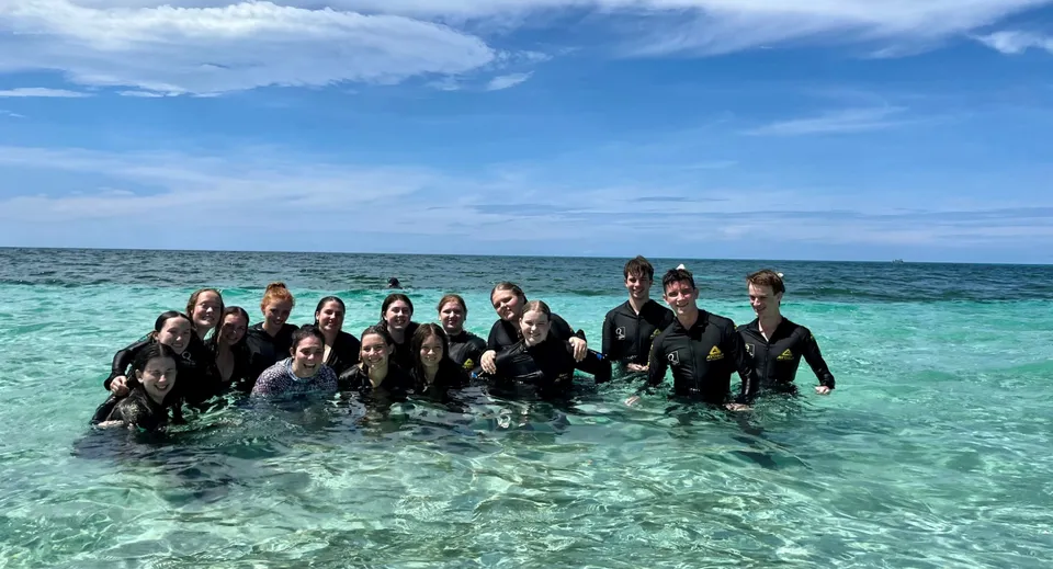 Un gruppo di studenti del Burpengary State Secondary College si trova in muta da sub nell'acqua bassa e turchese di una spiaggia tropicale.