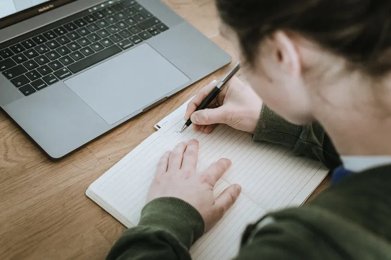 Person schreibt mit Kugelschreiber in ein Notizbuch neben einem Laptop