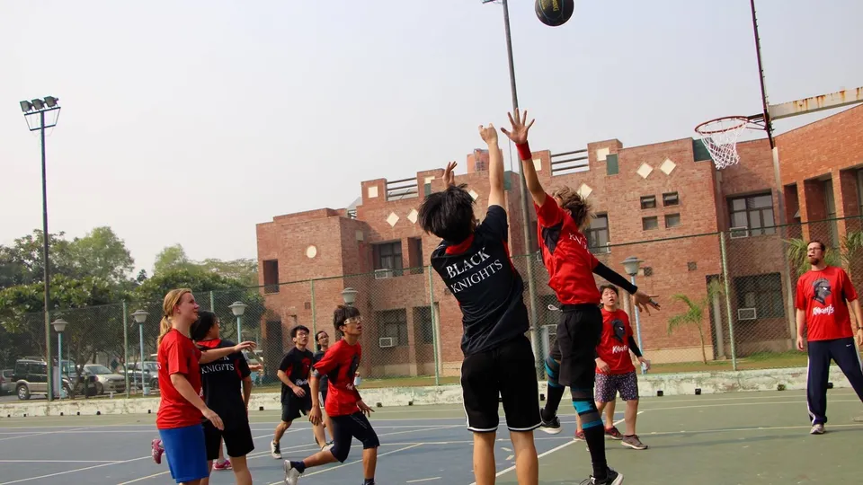 Studenti della Metro International Secondary Academy giocano a basket su un campo all'aperto davanti a edifici in mattoni.