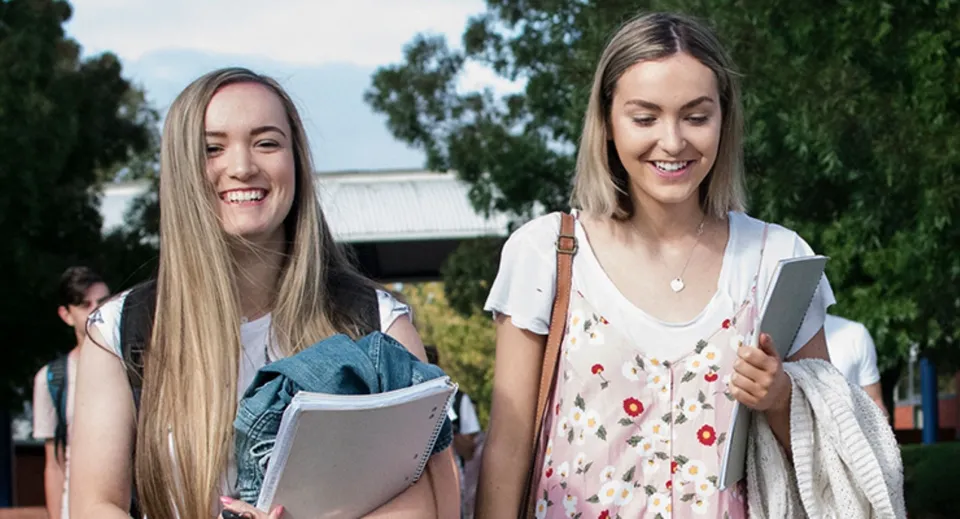 Due studentesse del Newstead College camminano sorridenti nel campus portando libri e raccoglitori.