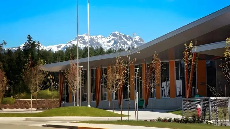 Flacher Schulbau mit auskragendem Dach und Fahnenmasten vor einer schneebedeckten Bergkette