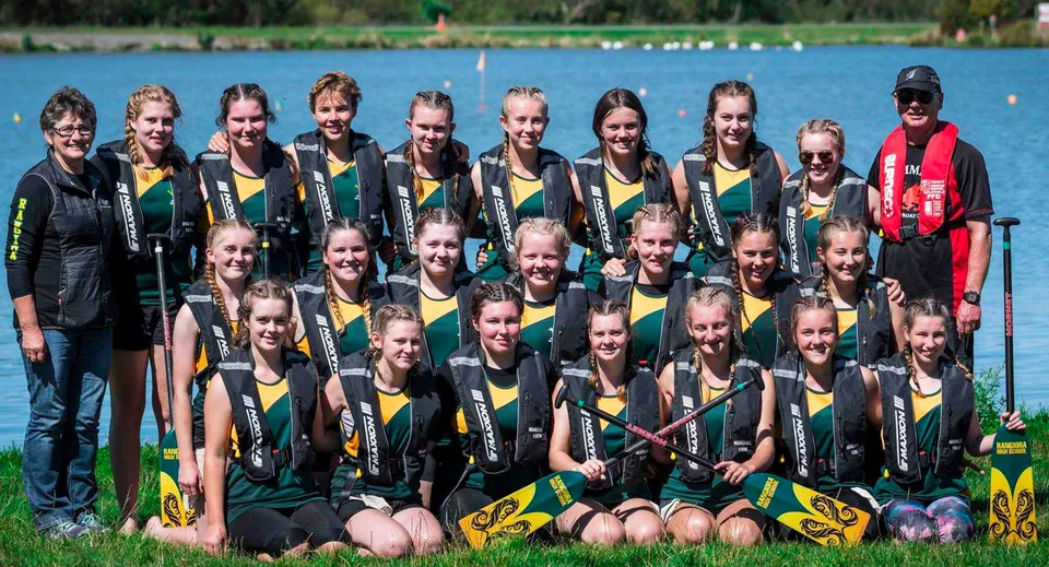 Un gruppo di giovani sportive in maglie verdi e gialle posa davanti a un lago con alberi nel campus della Rangiora High School.