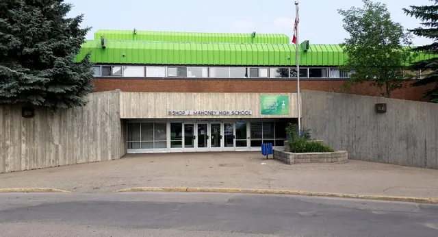 L'edificio moderno con il tetto verde si trova sull'area pavimentata della Bishop Mahoney High School, circondato da alberi e cespugli.