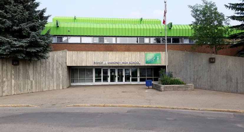 L'edificio moderno con il tetto verde si trova sull'area pavimentata della Bishop Mahoney High School, circondato da alberi e cespugli.