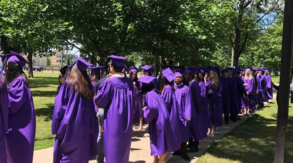Un gruppo di diplomati in toga viola e tocco cammina sul verde campus della Assumption College Catholic High School.