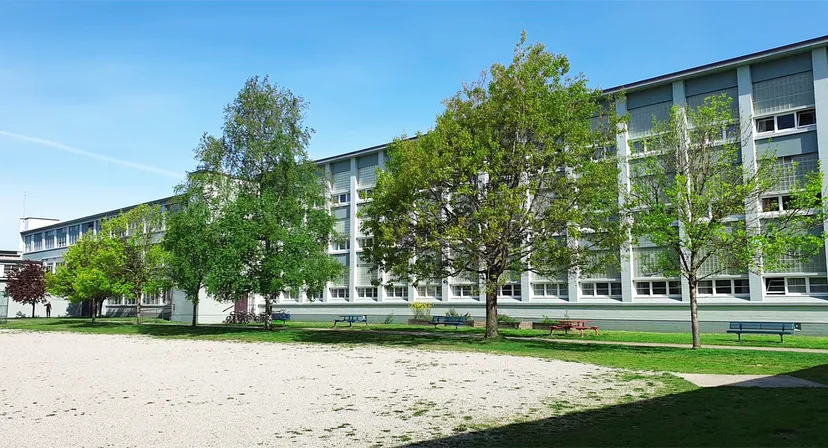 L'edificio scolastico a più piani della John Oliver Secondary School con alberi verdi e un cortile erboso sotto un cielo blu.