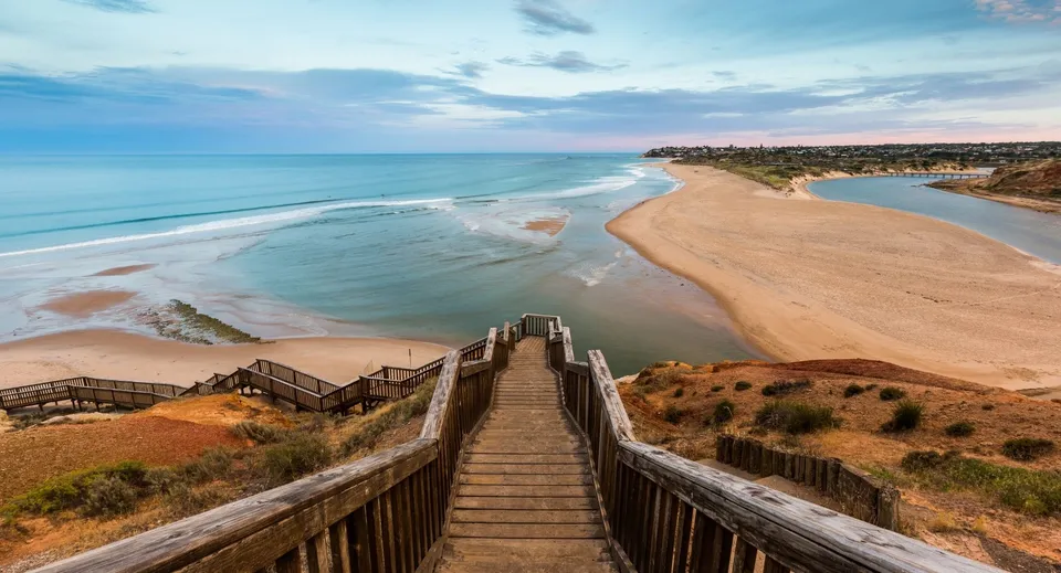 Una scala in legno conduce dal Mercedes College a un'ampia spiaggia di sabbia con acqua turchese e cielo nuvoloso.