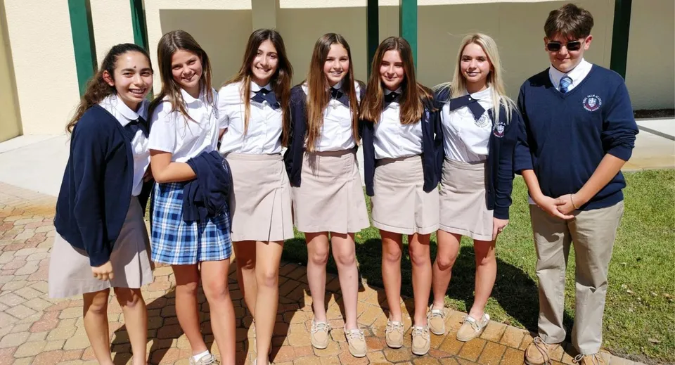 Un gruppo di studenti e studentesse della Royal Palm Academy si trova all'esterno, davanti a un edificio su una piazza pavimentata.