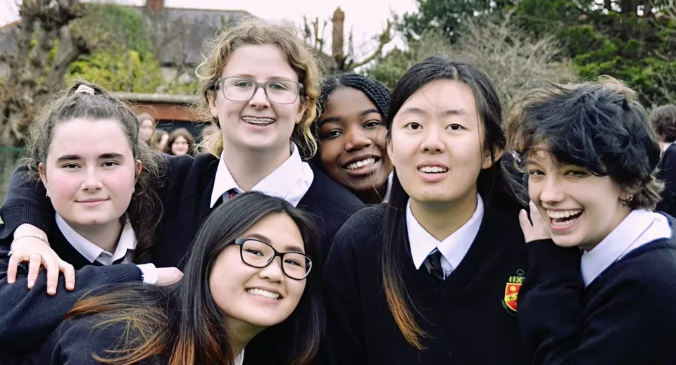 Un gruppo di studenti sorridenti di The High School è insieme all'aperto davanti ad alberi ed edifici.