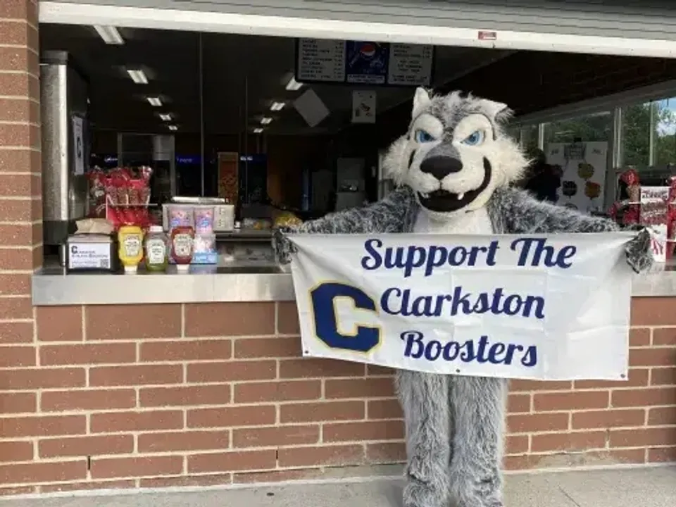 La mascotte della Clarkston High School regge uno striscione con la scritta Support The Clarkston Boosters davanti a un chiosco.
