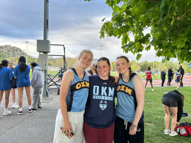 Tre studentesse in maglia OKM sorridono a bordo campo durante un evento di atletica in Canada