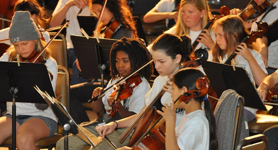 Un gruppo di studentesse della Salmo Secondary School suona diversi strumenti ad arco sul palco davanti a un pubblico.
