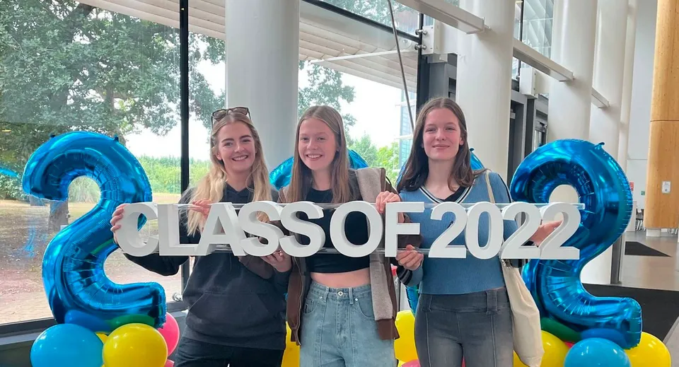 Tre studentesse tengono un grande cartello con CLASS OF 2022 davanti a un edificio del campus del Cheshire College South & West.