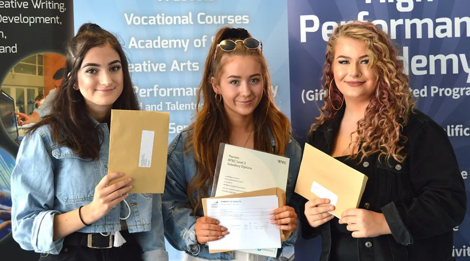 Tre studentesse mostrano i loro attestati davanti a uno sfondo informativo dell'Itchen Sixth Form College.