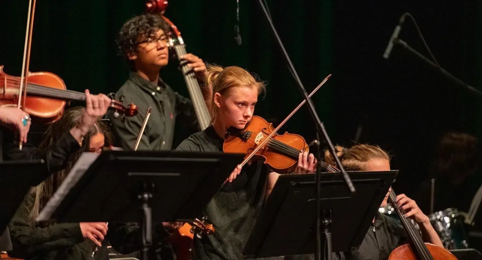 Un gruppo di musicisti della Frederick Gunn School si esibisce sul palco; in primo piano si vedono un violinista e un uomo con un microfono.