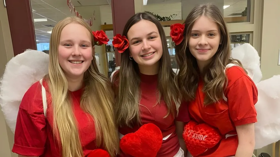 Tre studentesse della École secondaire Népisiguit indossano magliette rosse con motivi a cuore e sorridono in un ambiente interno.