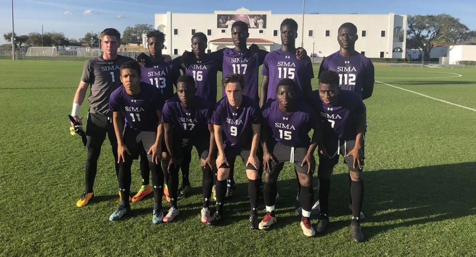 Un gruppo di calciatori con maglia viola si trova sul campo da calcio della Montverde Academy con un edificio sullo sfondo.