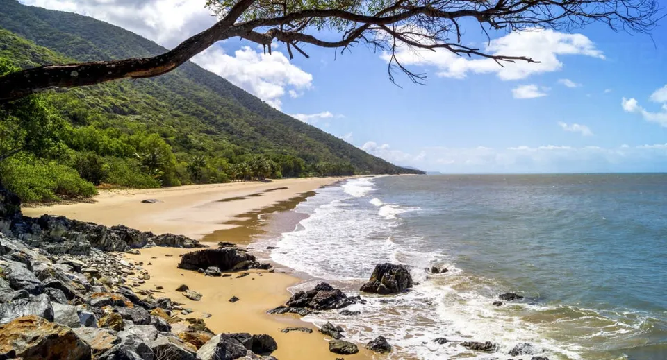 Un paesaggio costiero con spiaggia sabbiosa, rocce e montagne verdi, ripreso nei pressi della All Saints Anglican School.
