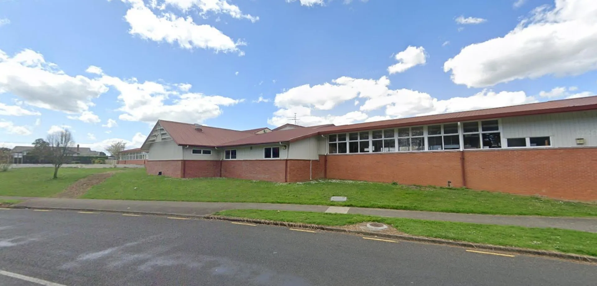 L'edificio in mattoni con tetto rosso si trova nel campus della Pukekohe High School, circondato da prati e una strada asfaltata.