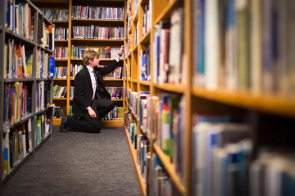 Una persona con un cappotto nero è in piedi nel corridoio stretto tra alti scaffali di libri nella biblioteca del Campbell College.