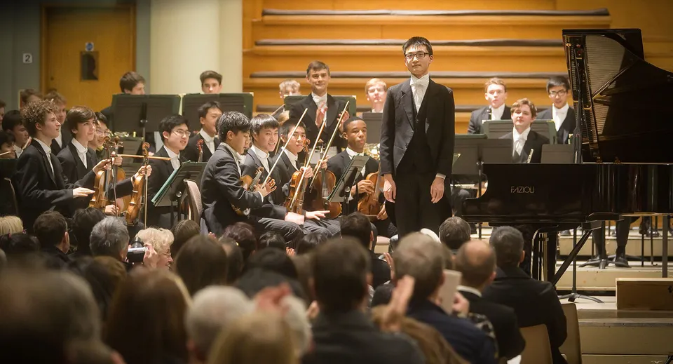 Un'orchestra dell'Eton College si esibisce sul palco con un direttore d'orchestra e un pianista al pianoforte a coda.
