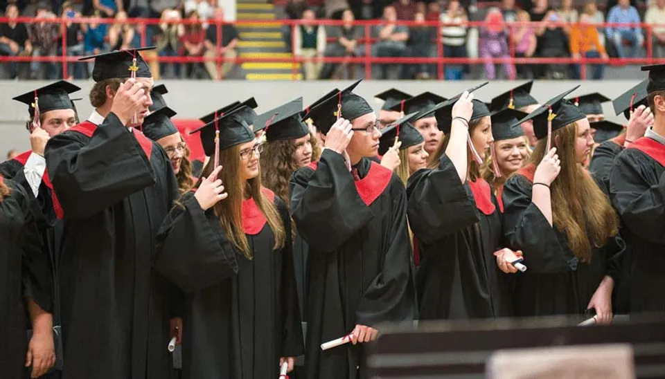 Un gruppo di diplomati della Lambton Central Collegiate & Vocational School è in piedi con toghe nere davanti a una parete colorata con bandiere.