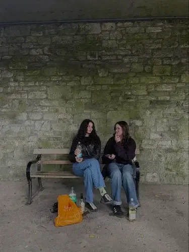 Dos chicas charlando sentadas en un banco bajo un puente de piedra