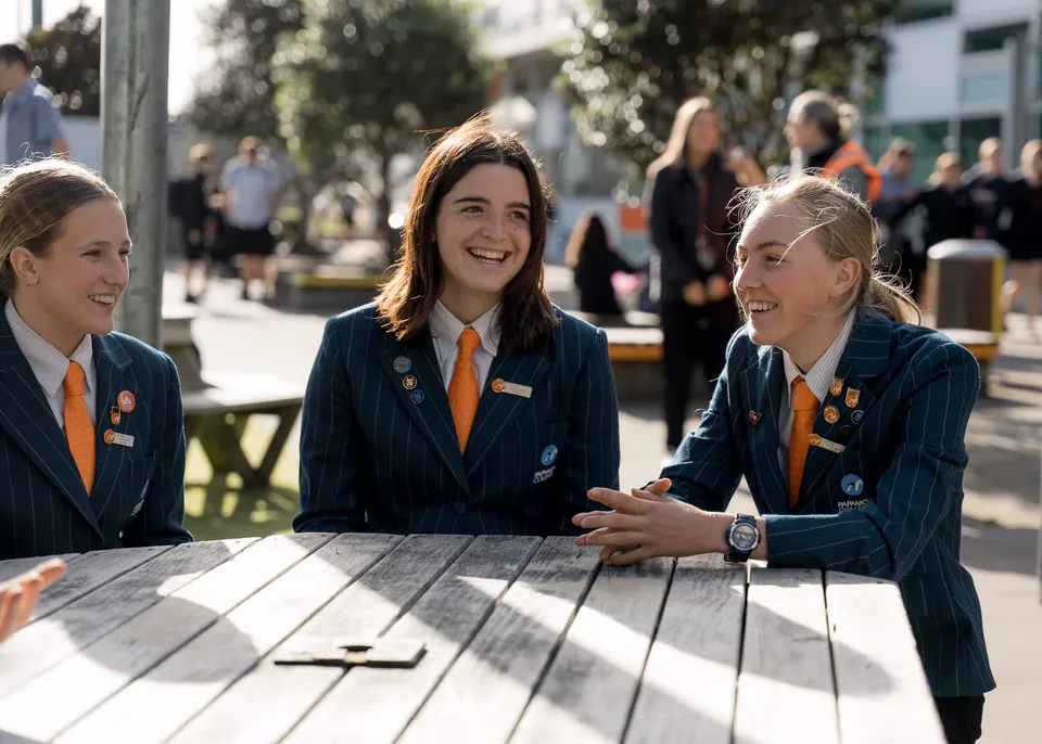 Tre studentesse in uniforme scolastica sedute a un tavolo di legno chiacchierano nel campus del Papamoa College.