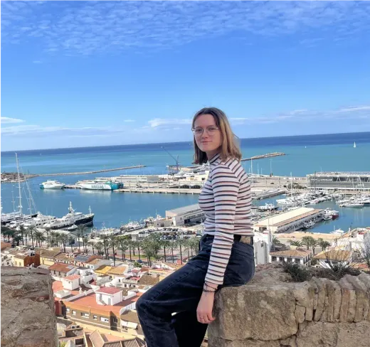 Mariea sitzt auf einer Mauer über dem Yachthafen, im Hintergrund das Meer, in Spanien