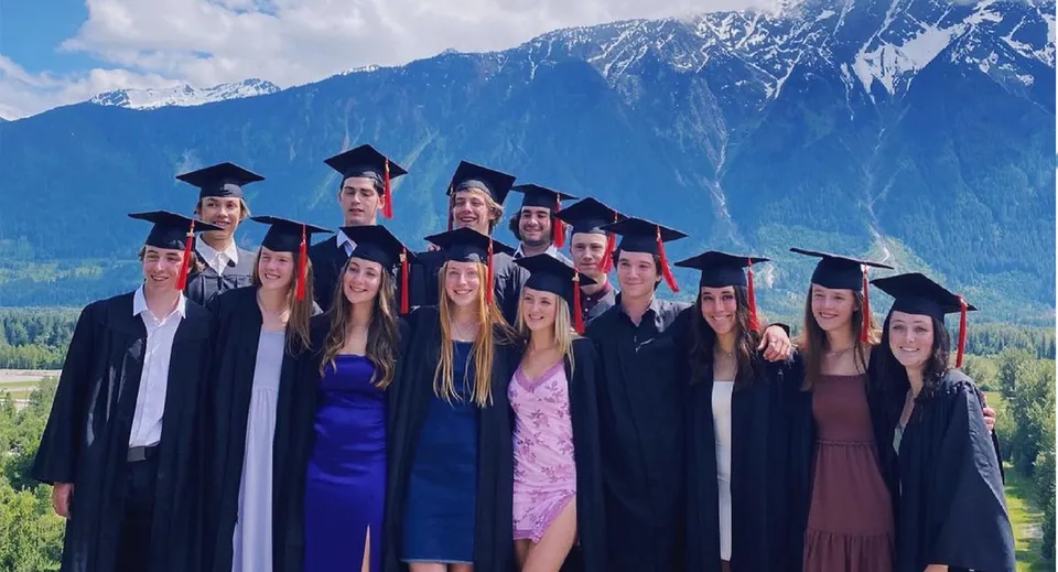Un gruppo di diplomati in abito accademico si trova davanti a un paesaggio montano nel campus della Pemberton Secondary School.