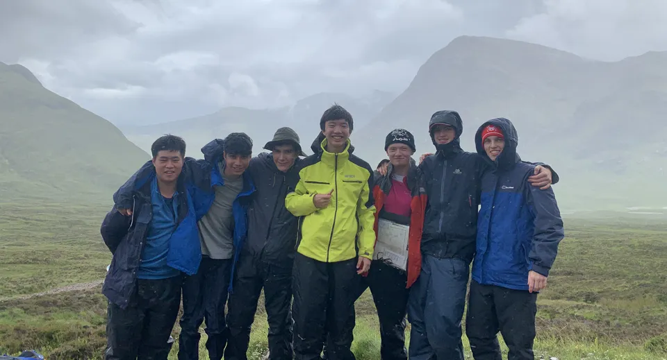 Un gruppo di studenti di Ashville College in abbigliamento da pioggia davanti a un paesaggio montuoso.