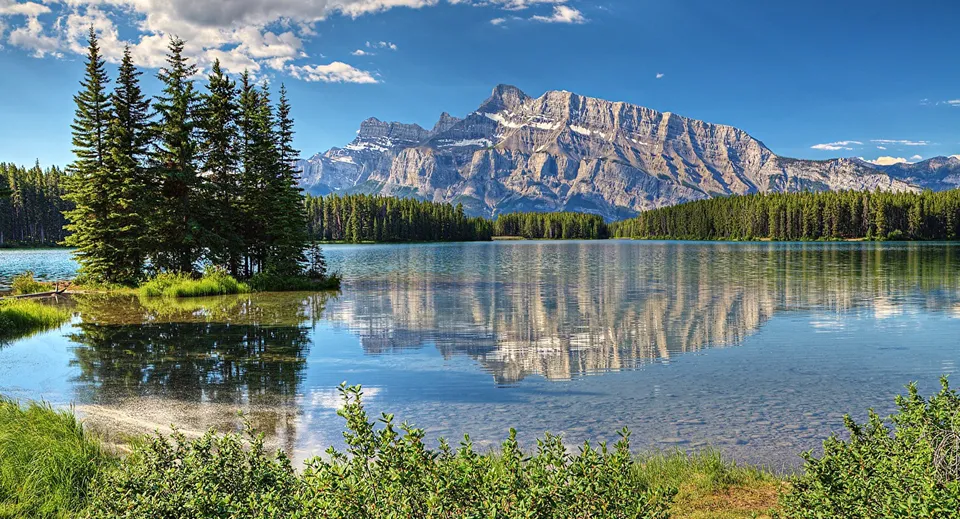Un lago calmo riflette una foresta di pini e una grande montagna rocciosa sotto un cielo sereno.