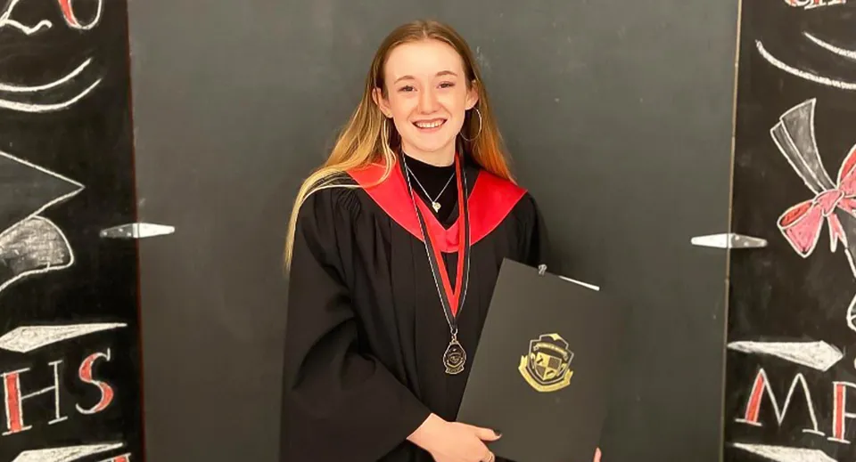 Una giovane donna con la toga di laurea sta in piedi davanti a una lavagna con disegni nel campus della Montague Regional High School.