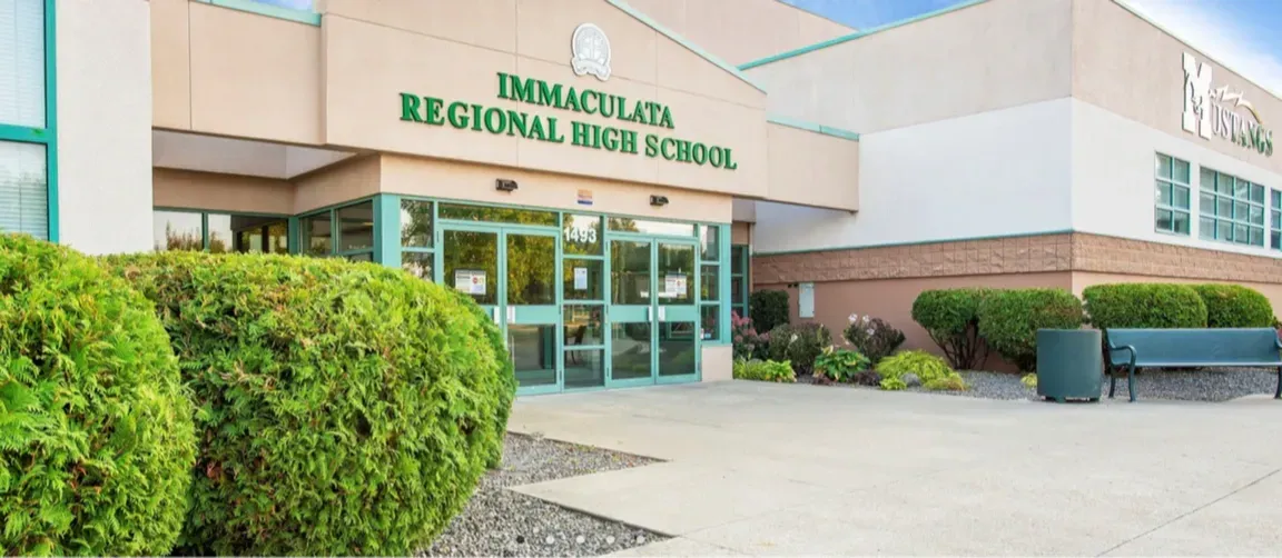 Entrada de un edificio escolar con el rótulo Immaculata Regional High School, arbustos y un banco delante