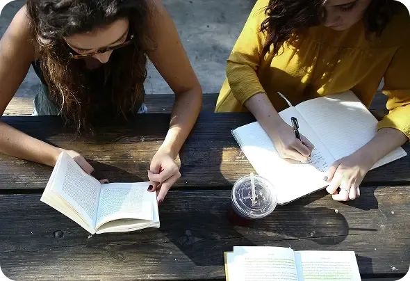 Due studentesse studiano all'aperto a un tavolo di legno, una legge un libro e l'altra prende appunti