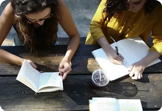 Due studentesse studiano all'aperto a un tavolo di legno, una legge un libro e l'altra prende appunti