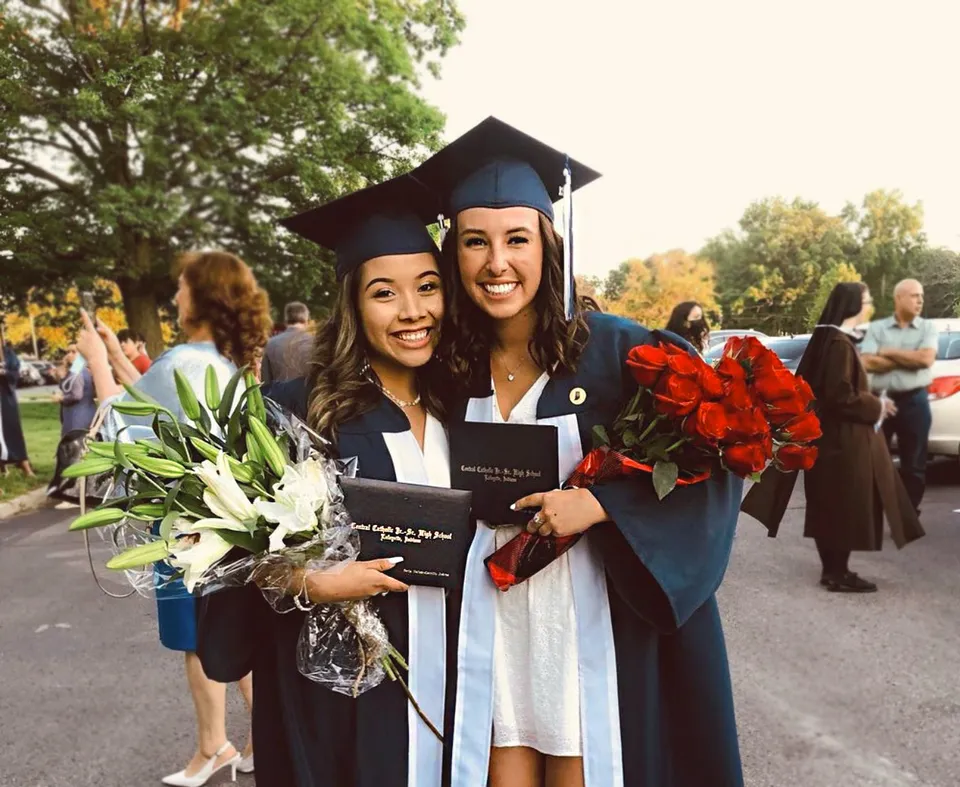 Due studentesse della Central Catholic Jr.-Sr. High School in abito da diploma tengono dei fiori e sorridono con altri diplomati sullo sfondo.