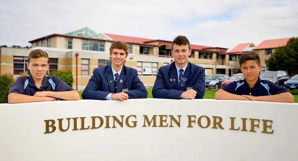 Quattro studenti della King's High School siedono davanti al cartello 'BUILDING MEN FOR LIFE' con l'edificio scolastico sullo sfondo.
