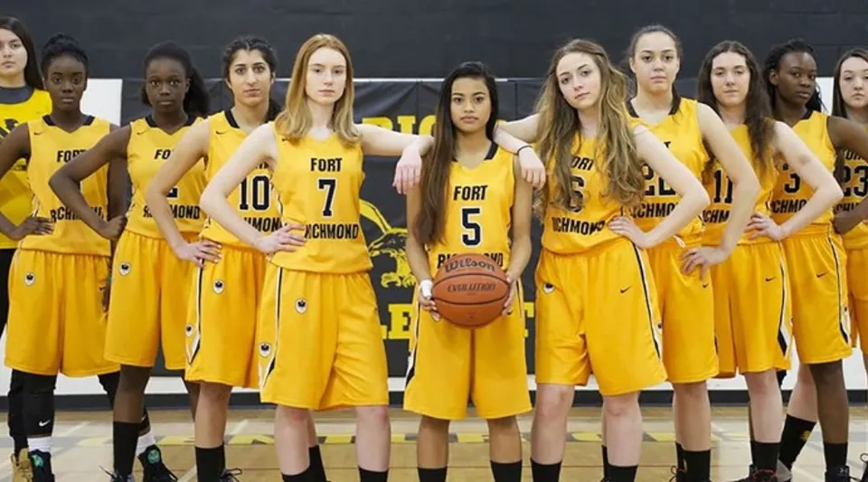 Un gruppo di giovani donne in maglia da basket gialla sta insieme, una di loro tiene un pallone da basket, foto scattata nel campus del Fort Richmond Collegiate.